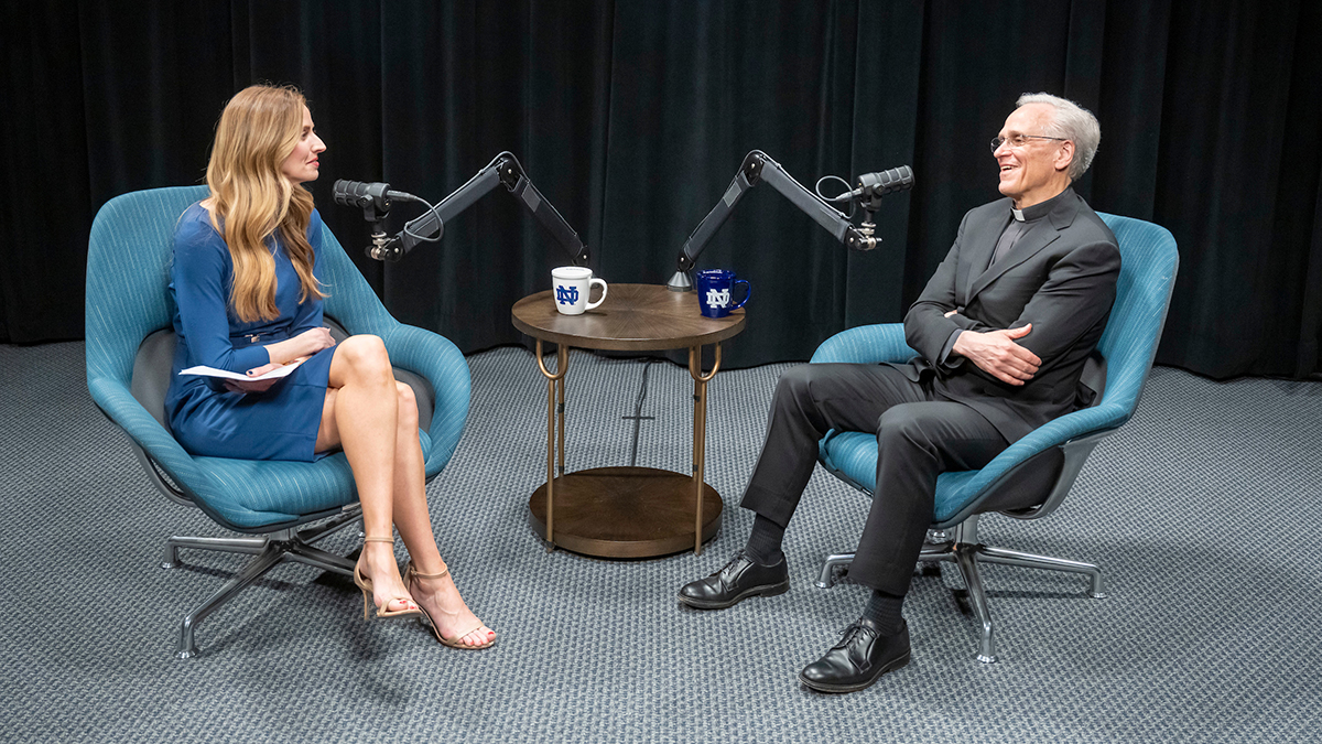 A blonde female in a blue dress sits smiling across from a smiling man with grey hair wearing a dark suit and a priest's collar. They are separated by a small circular table and two microphones. On the table are two Notre Dame coffee mugs: one white, one blue. Behind them is a black curtain backdrop.