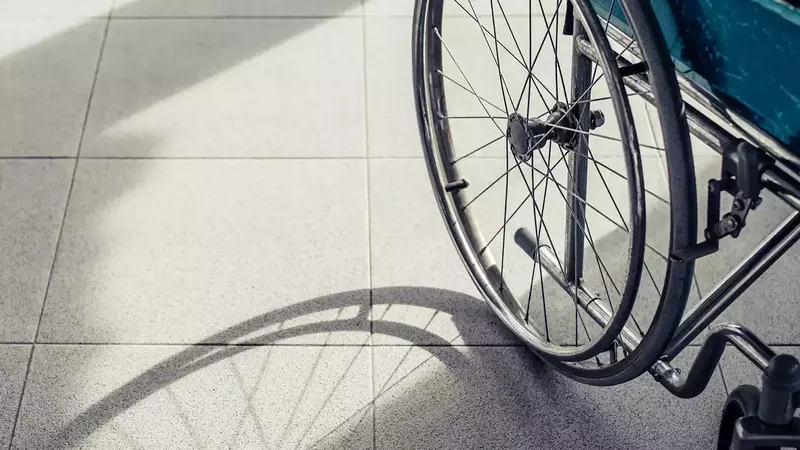 A wheelchair sits on a sunny expanse of speckled tile, its back to the camera, casting a strong shadow that stretches toward the viewer.