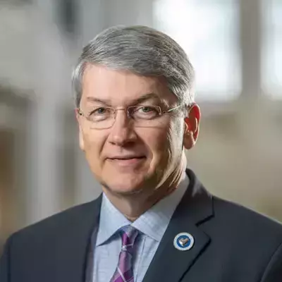 A white man with short gray hair and glasses. He wears a dark suit jacket, white shirt, and purple patterned tie. A small, round Notre Dame pin is affixed to his lapel.