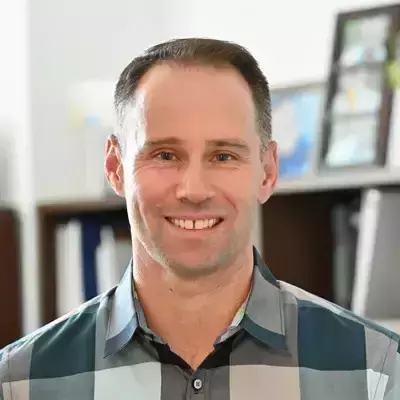 A white man with short brown hair, smiling, wearing a gray and green plaid button-down shirt. He is positioned in front of a bookshelf slightly out of focus.