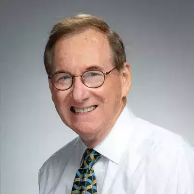 A white man with light brown hair and glasses, wearing a white collared shirt and patterned tie. He is smiling at the camera against a plain gray background.