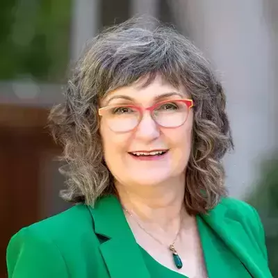 A white woman with short, gray, curly hair and red-rimmed glasses. She smiles warmly while wearing an emerald green blazer over an emerald top. A teardrop-shaped green pendant necklace hangs around her neck. A blurred background hints at an outdoor setting.
