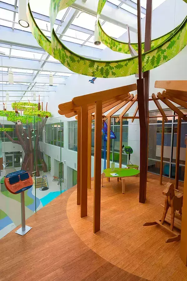 The atrium area of the NICU unit has a whimsical, indoor treehouse play area with light wood framing, flooring, and furniture. In the background, another tree sculpture and play area are visible through glass partitions.
