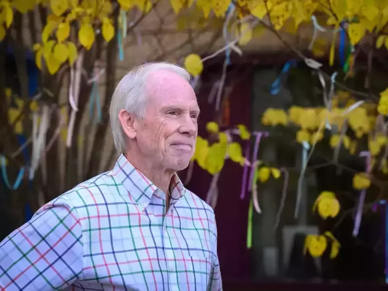 An white man with short white hair wears a light blue, red, and green plaid button-down shirt. He stands slightly angled to the right of the frame, looking pensively in that direction. Behind him, branches with yellow fall leaves are decorated with colorful ribbons.