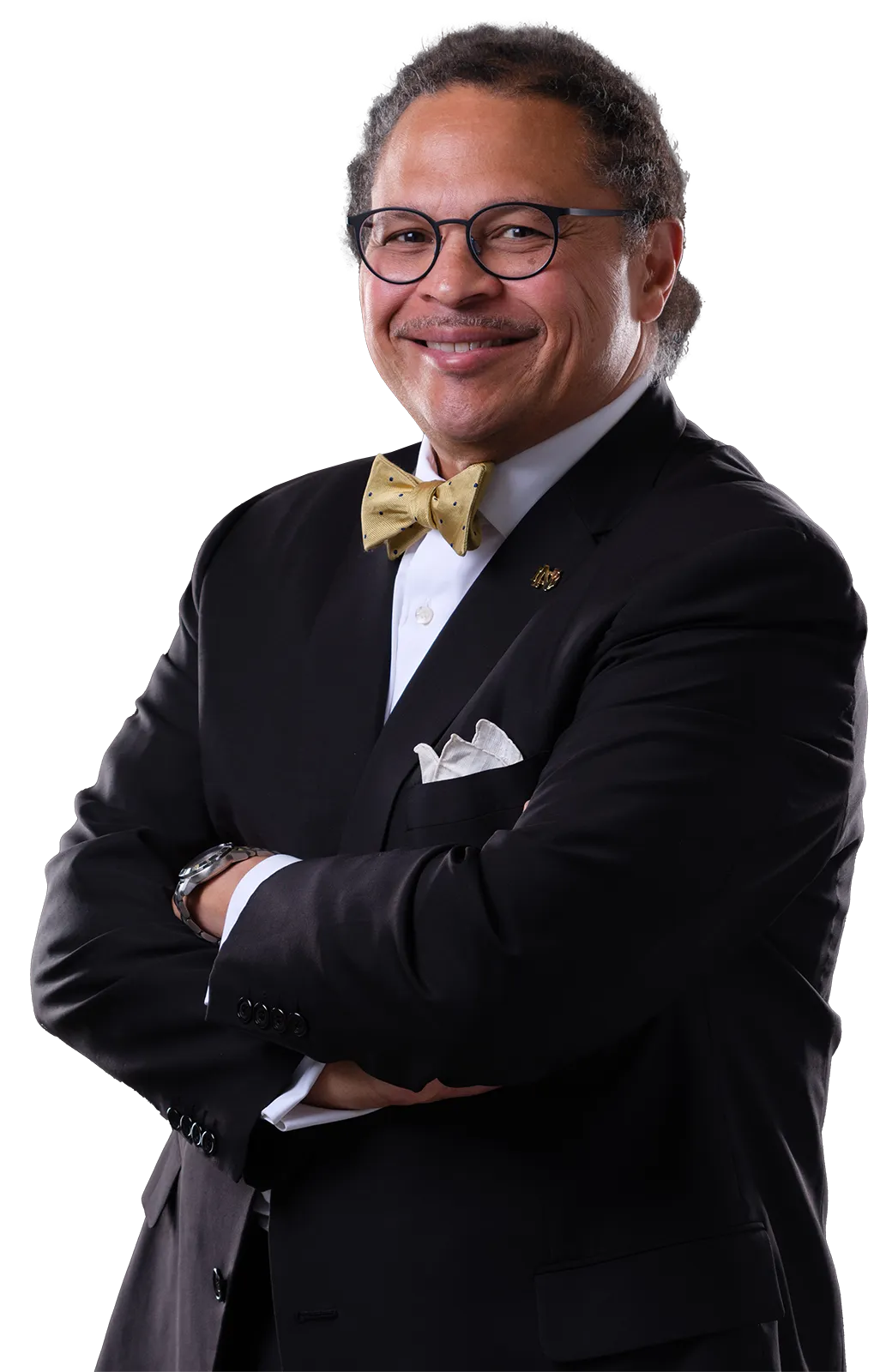 A man smiles at the camera with his arms crossed. He wears a dark suit, white shirt, gold patterned bow tie, and a white pocket square. A small, gold Notre Dame pin is visible on his lapel.