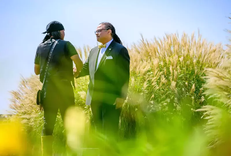 Two individuals shake hands amidst tall, ornamental grasses on a sunny day. One person wears a black bandana, shirt, and pants, while the other wears a suit with a yellow bow tie.