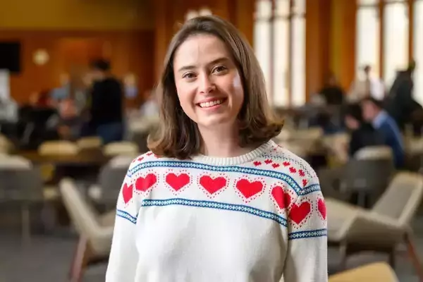 A white woman smiles at the camera in a common area inside a University of Notre Dame building. She is wearing a white sweater with a red and blue heart pattern across the chest and shoulders. The background is slightly blurred, showing other students and seating.