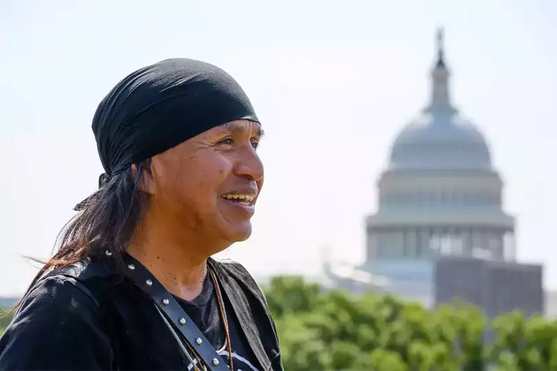 A Native American man wearing a black bandana and dark shirt smiles while looking off to the side. The U.S. Capitol Building is visible in the background, slightly blurred by the focus on the person in the foreground. Lush green trees line the bottom of the frame.