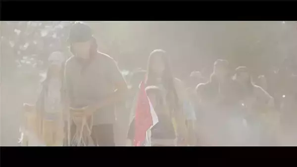 A hazy, dreamlike image of people walking through a thick fog. Figures in the foreground are slightly more visible, including a person in a dark hat and jacket and another with a reddish garment. The background is filled with blurred shapes of other individuals, making the scene appear mysterious and ethereal.