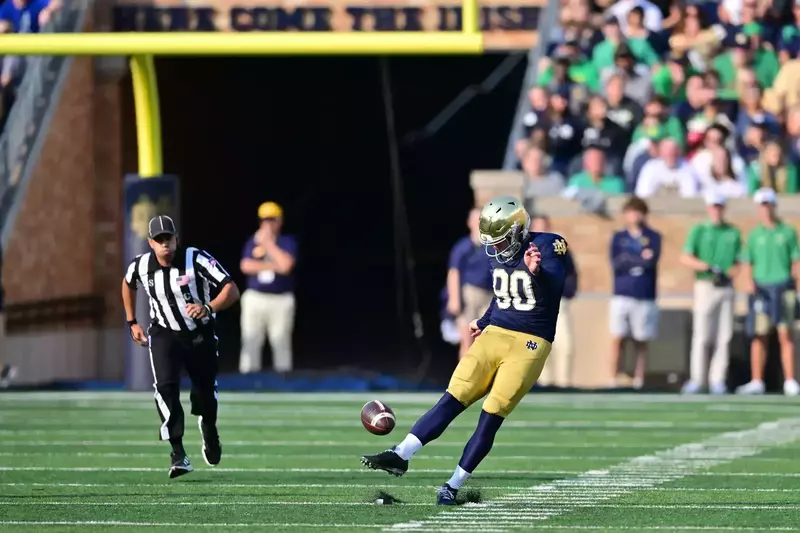 Football player Eric Goins, number 90, kicks the ball during a game. A referee in black and white stripes runs towards the ball in the foreground.
