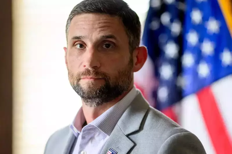 Headshot of a white man with short, dark hair, a beard, and a light gray blazer. An American flag is slightly blurred in the background.
