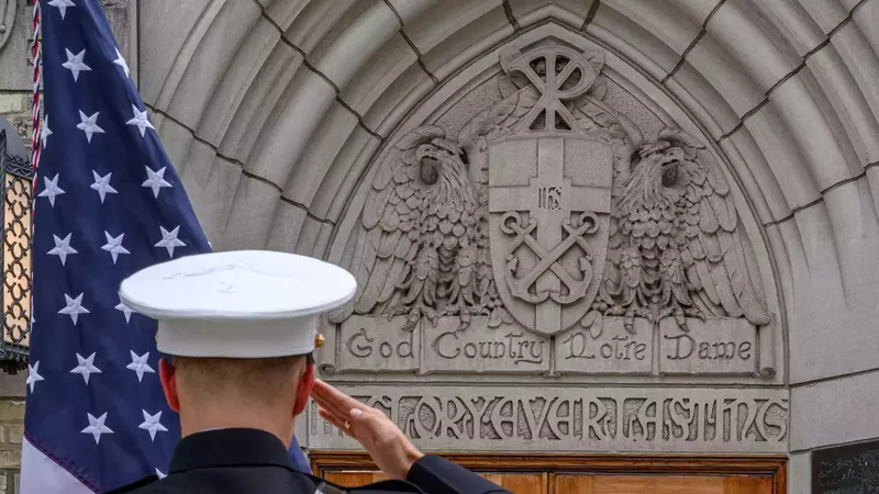 A U.S. service member in a white cap salutes the American flag in front of a stone archway at the University of Notre Dame. The arch is decorated with two sculpted eagles flanking the university's seal, which includes crossed anchors and the Chi Rho symbol. Below the seal are the words "God, Country, Notre Dame."