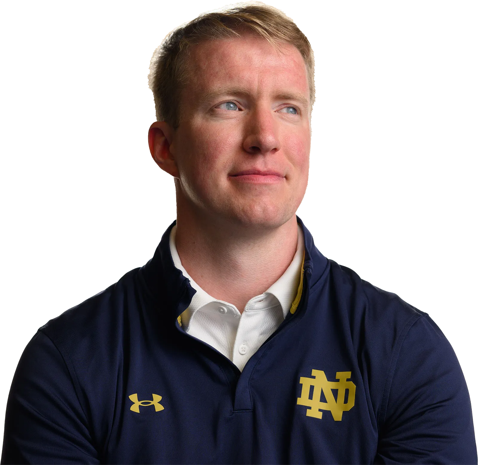 A man wearing a navy blue polo shirt with the gold "ND" logo, representing the University of Notre Dame, looks off to the side with a thoughtful expression.	He wears a white collared shirt underneath the polo shirt.	An Under Armour logo is visible on the left side of the polo shirt.