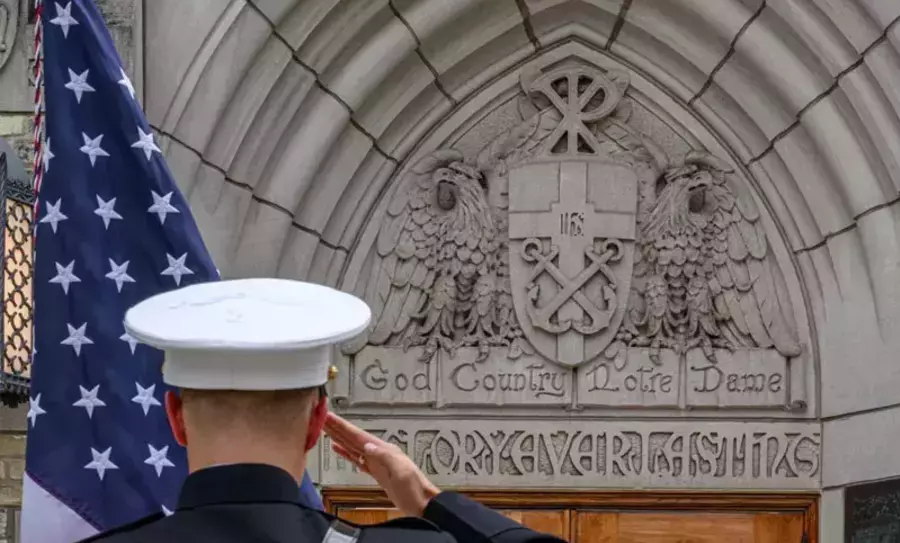 A U.S. serviceman in a white dress uniform salutes in front of a stone archway at the University of Notre Dame. The arch is adorned with an eagle, a crest, and the inscription "God, Country, Notre Dame." An American flag is positioned to the serviceman's left.