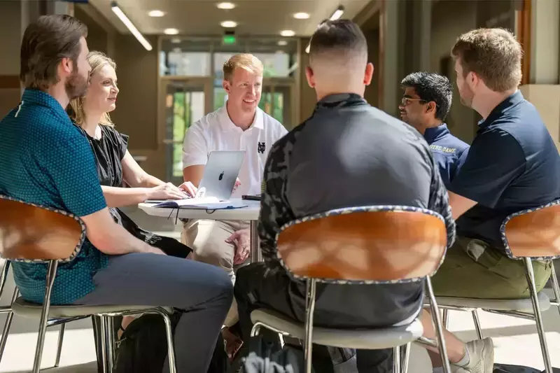 A group of Notre Dame students and faculty sit around a small table, engaged in a discussion. A laptop and notebook rest on the table, suggesting a collaborative project or meeting.