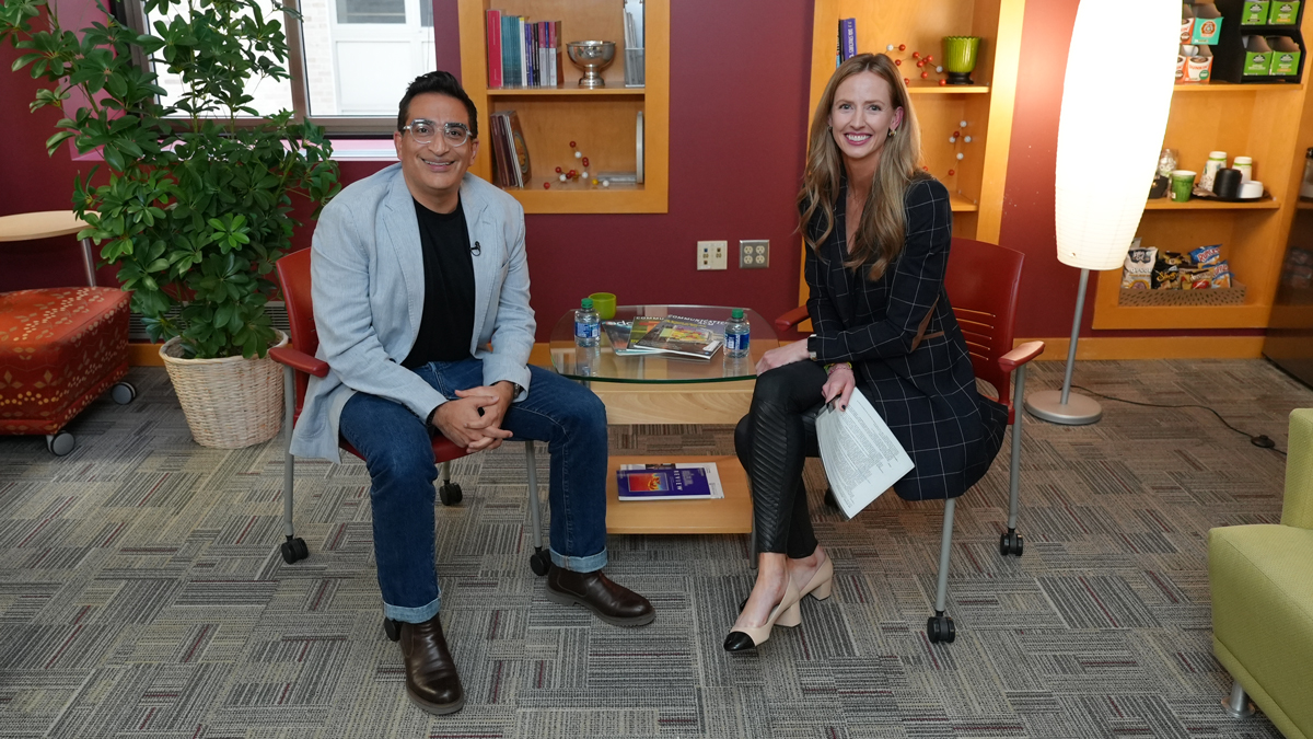 An Indian man and a white woman sit facing the camera in red rolling chairs around a small glass table. The room has red walls, a patterned carpet, potted plants, and a bookshelf visible in the background. They appear to be in a casual conversation or interview setting.