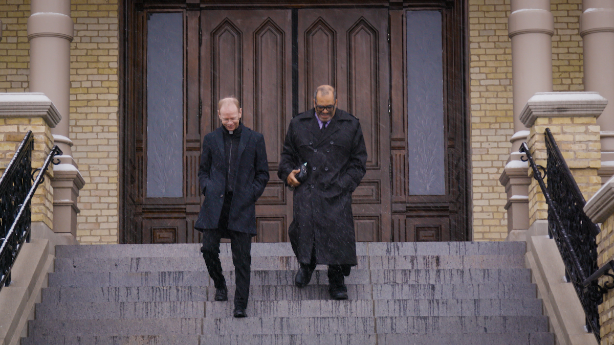 Two men descend the steps of a light brick building with a large wooden door as snow falls. The man on the left is white and wears a clerical collar and black coat. The man on the right is Black and wears a black trench coat.