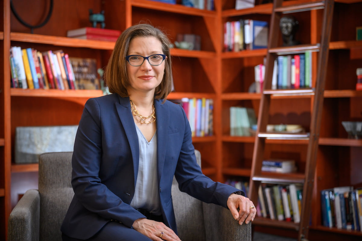 A woman with shoulder-length brown hair and glasses sits in a gray chair in front of bookshelves. She wears a navy blue suit jacket over a light blue v-neck shirt and a gold chain necklace. She is posed with her hands resting on the arms of the chair.