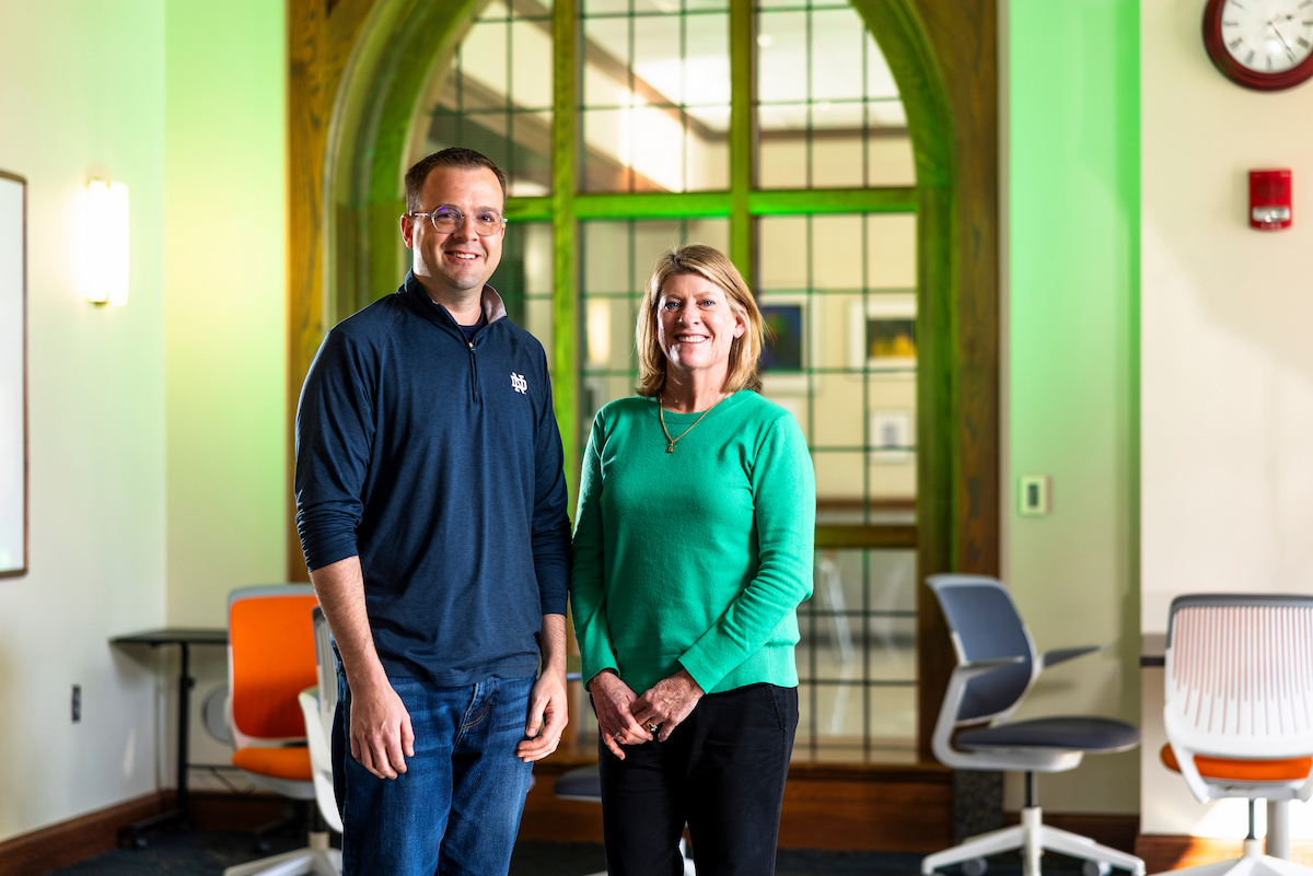 A white man and woman stand smiling in a lounge area. The man wears a navy blue quarter-zip with the ND logo and jeans. The woman wears a green sweater and black pants. They stand before a large, arched, multi-paned window with green lighting.