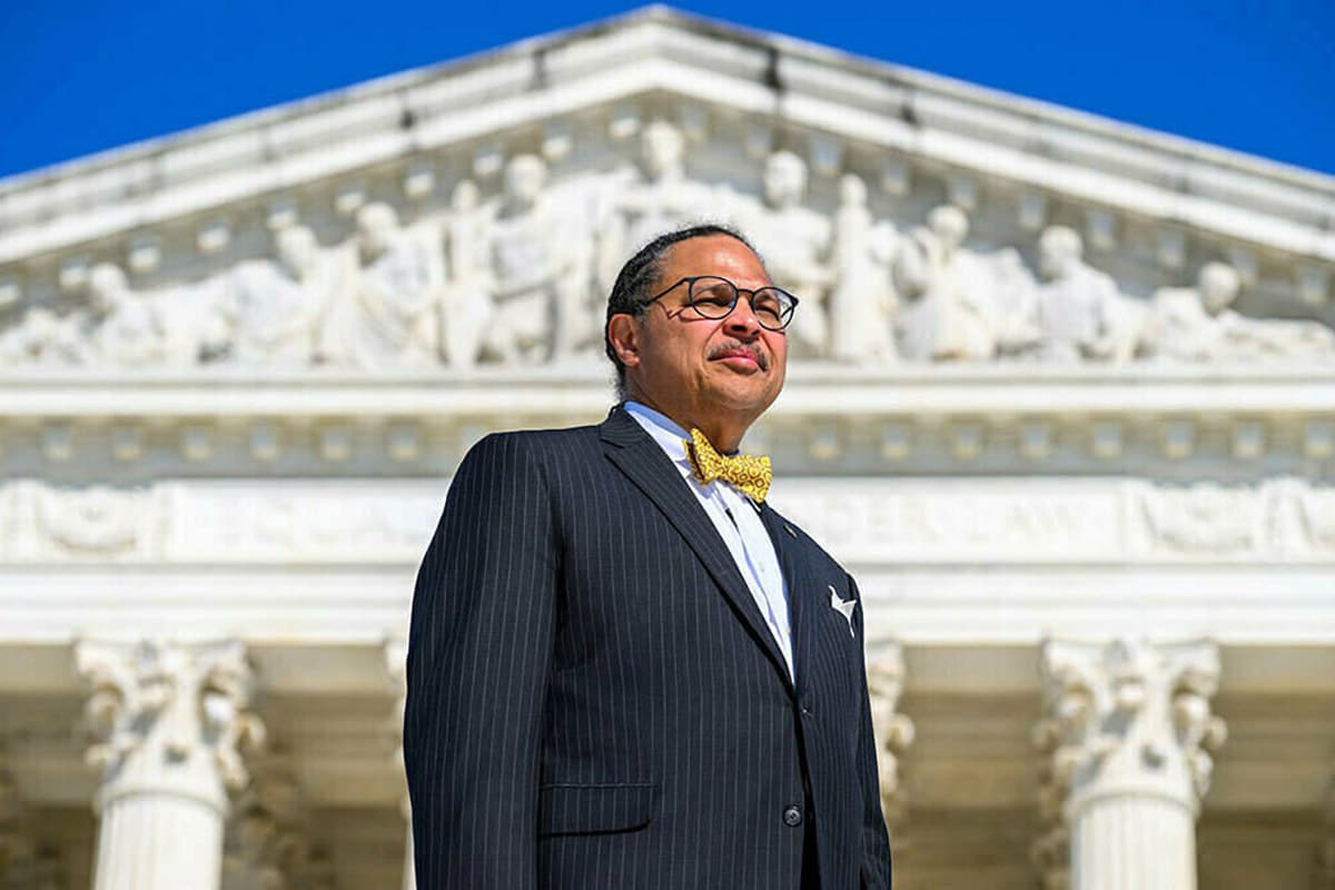 A man in a dark pinstripe suit and gold bow tie stands before the U.S. Supreme Court building. He wears glasses and looks upward.