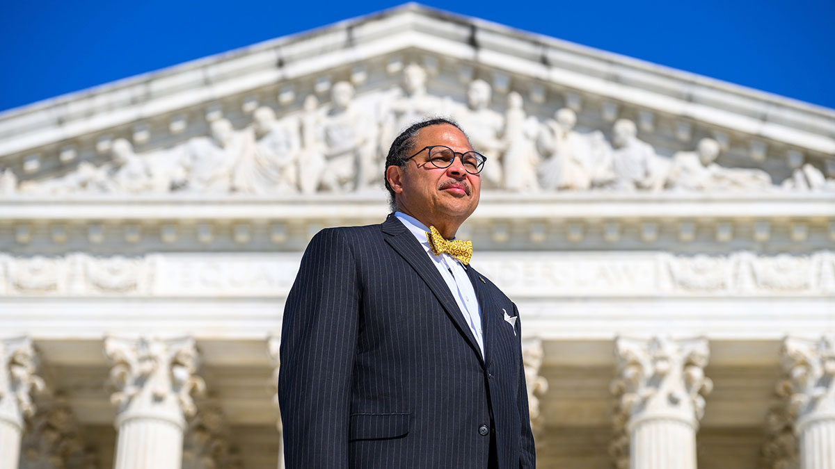 A man in a dark pinstripe suit and gold bow tie stands before the U.S. Supreme Court building. He wears glasses and looks upward.