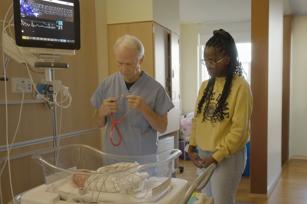 A doctor in blue scrubs holds a red stethoscope, preparing to examine a newborn lying in a hospital bassinet.  A young woman wearing a yellow Notre Dame sweatshirt stands beside them.