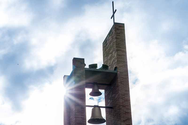 Sunlight shines behind a brick and concrete bell tower with two bells hanging beneath a cross. 