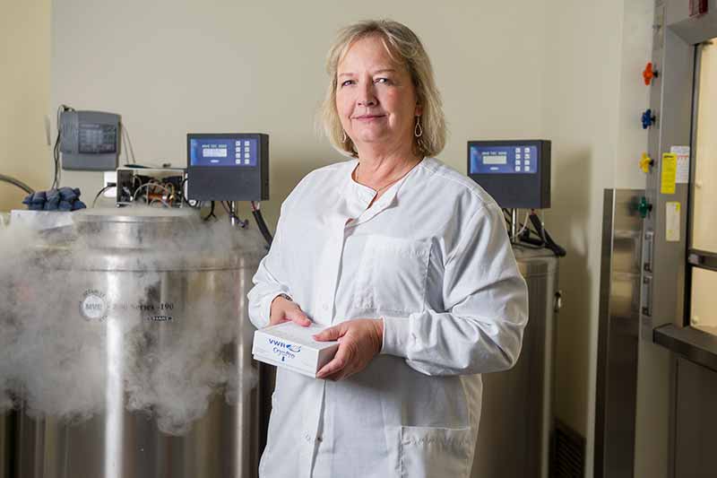 A woman in a white lab coat stands in a lab, holding a white box, with cryogenic storage tanks behind her.