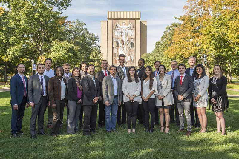 A diverse group of smiling students poses for a photo on the lawn in front of the Word of Life mural on Hesburgh Library.