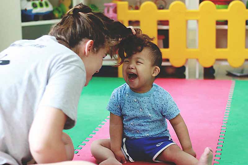 A smiling woman looks down at a laughing toddler with messy hair, who is sitting on a colorful foam mat.