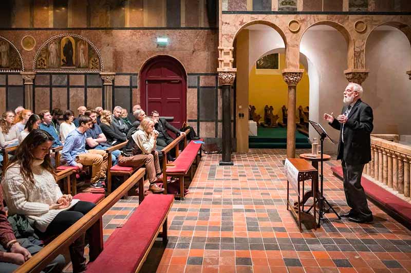 An older man with a white beard speaks to a seated audience in a hall with decorative columns. 