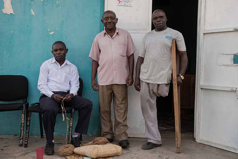 Three men stand outside a light blue building; one man is seated, and two are standing.