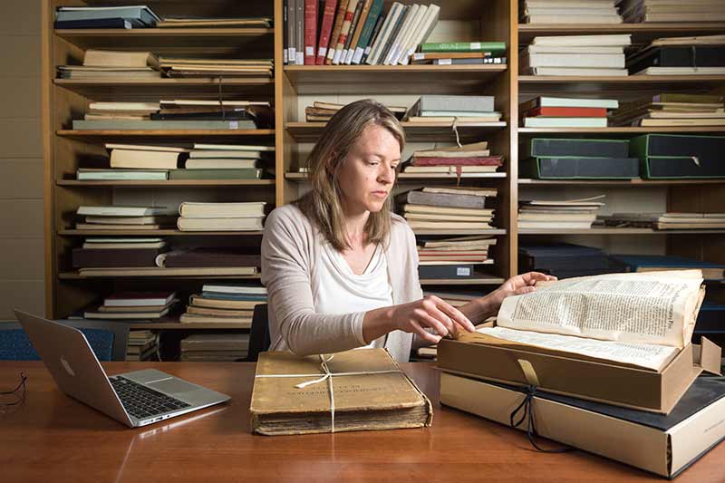 A woman sits at a desk with a laptop, looking through an archival document in a folder. 
