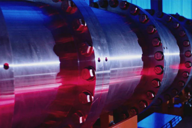 A close-up of a large, metallic industrial pipe with a series of bolted flanges and a blue and red light reflecting off the surface.