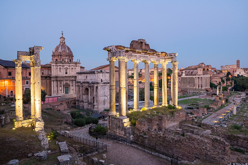 The ruins of the Roman Forum are lit up at dusk, with ancient columns and St. Peter's Basilica visible in the background. 