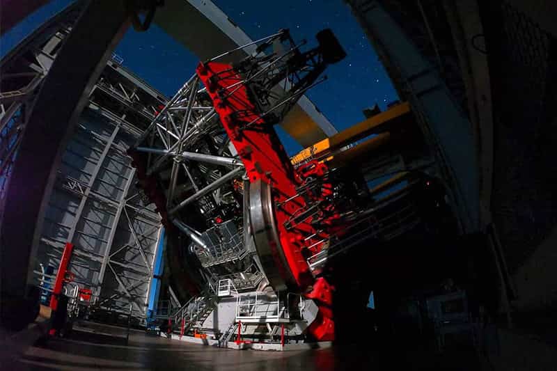An interior view of an observatory shows a large red telescope pointing toward a starry night sky. 