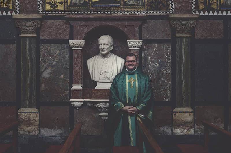 A smiling priest in a green vestment stands in front of a white bust of a man in the Basilica of the Sacred Heart.