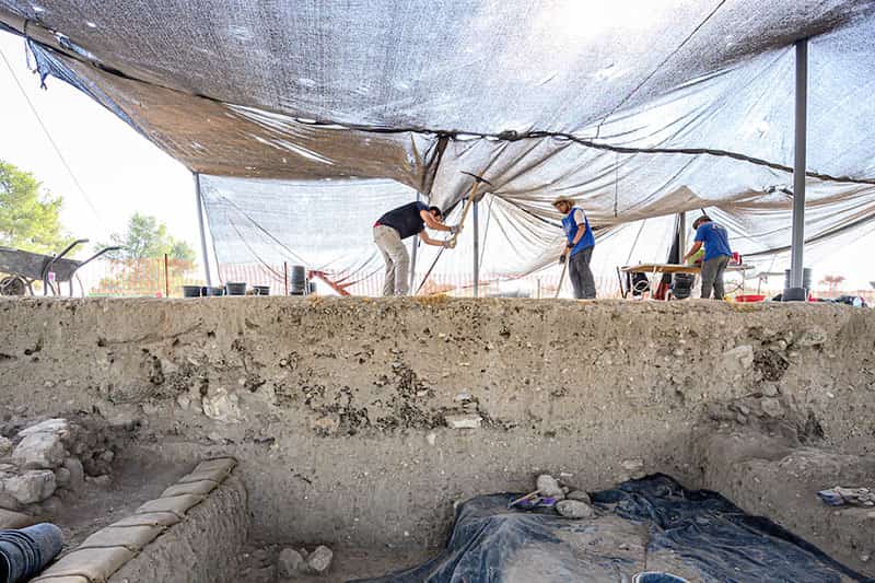 Archeologists work under a large tarp at an excavation site.