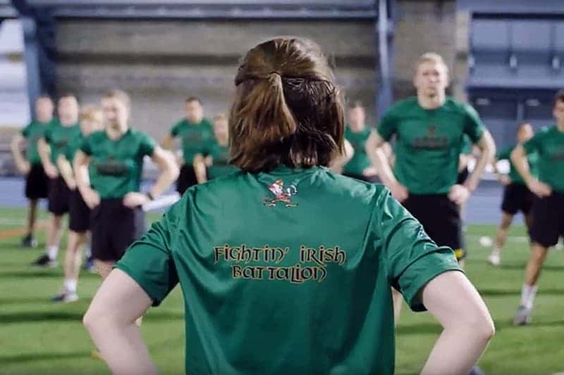 A person in a green shirt with &lsquo;FIGHTIN IRISH BATTALION&rsquo;on the back stands facing a group of people on a field.