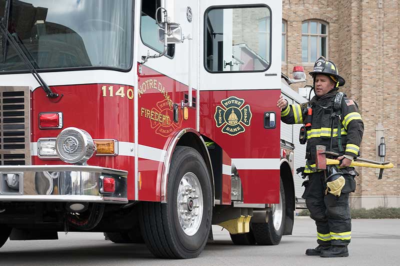 A Notre Dame firefighter in full gear with an axe stands next to a fire truck.