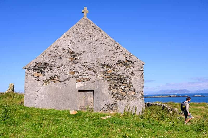 An old stone chapel with a cross on the roof stands on a green hill overlooking the blue ocean with small islands. 
