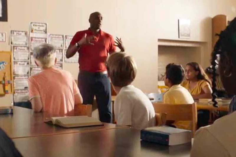 A man in a red polo shirt stands and gestures while speaking to a diverse group of adults sitting at a table in a classroom.