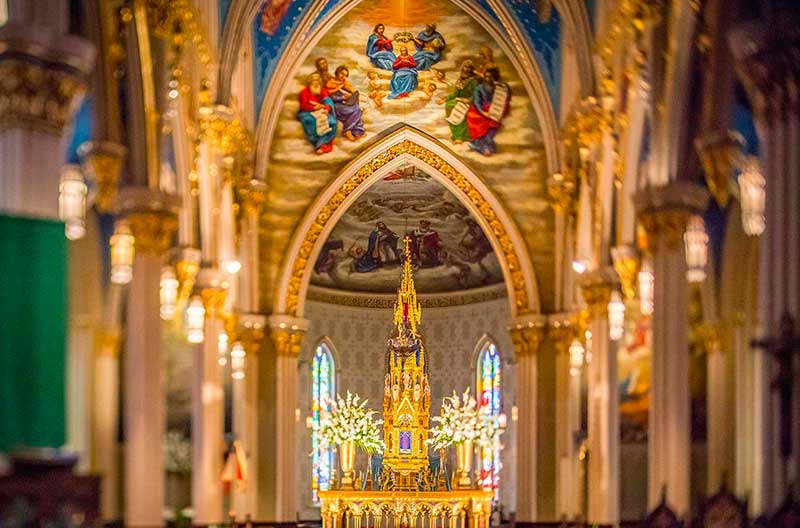 An ornate gilded altar inside the Basilica of the Sacred Heart at Notre Dame is surrounded by arches and murals on the ceiling.