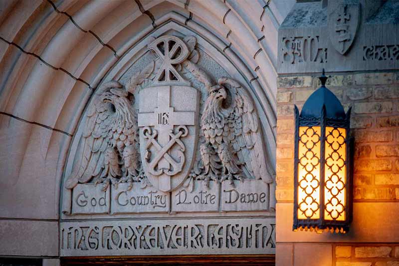 A Notre Dame coat of arms carved into stone with the motto &lsquo;God, Country, Notre Dame&rsquo; is illuminated by a decorative lantern.