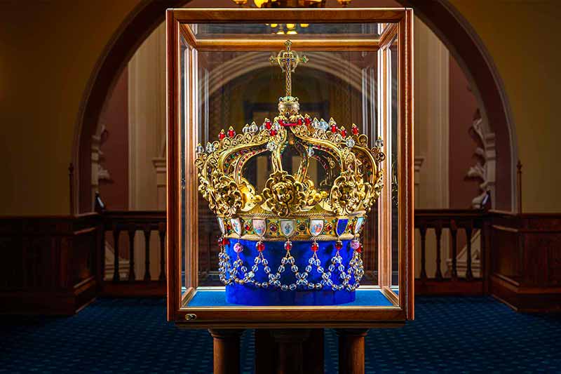 The ornate, golden crown of the statue of Mary atop the Golden Dome, with red jewels and a blue velvet base, displayed inside a glass case.