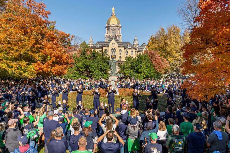 The Notre Dame Band of the Fighting Irish plays for a large crowd in The Fieldhouse Mall with the Main Building and Hesburgh Library in the background during a sunny fall day.