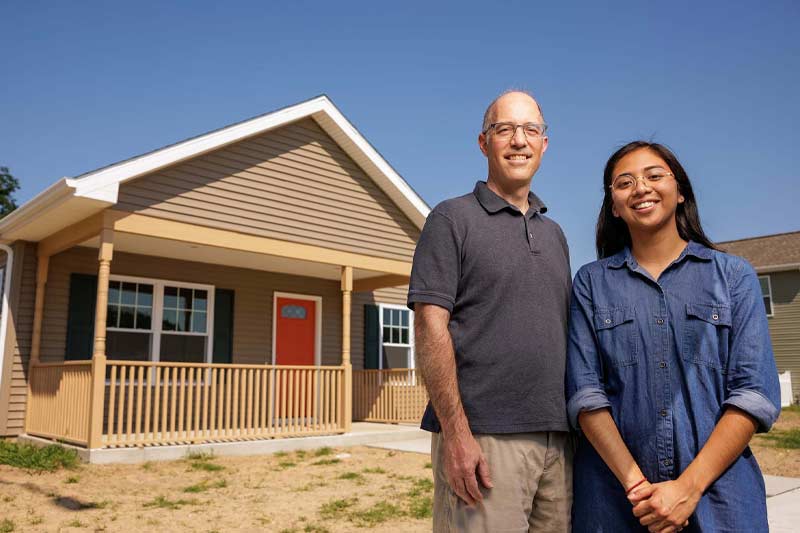 A smiling man and woman stand in front of a new house with tan siding and a red front door.