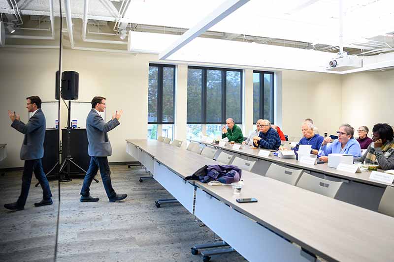 A man in a suit jacket gestures while giving a presentation to a seated audience at a long table in a conference room.