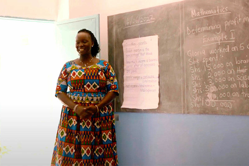 A smiling woman wearing a colorful patterned dress stands next to a chalkboard with math problems and a paper note.