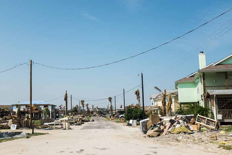 A street in a residential area is covered with debris and damaged belongings, with some houses showing visible damage. 